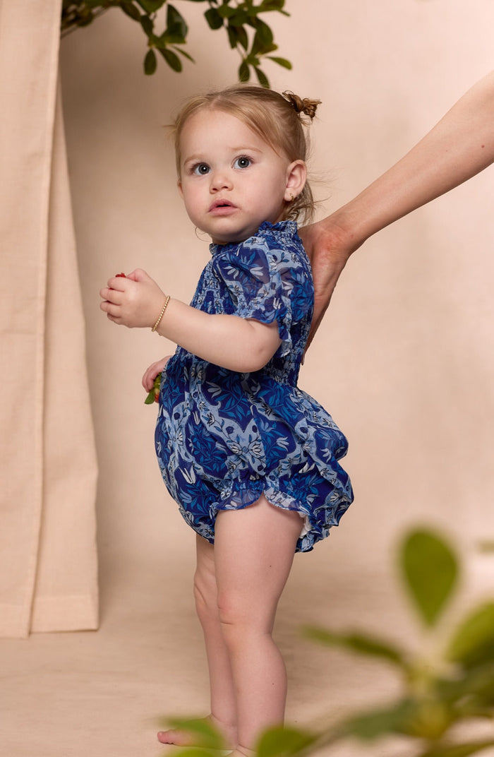 A toddler wearing the Ivy City Co Baby Cordelia Romper in Trellis Foulard, featuring a smocked bodice and blue floral print, stands sideways and looks up. An adult hand gently supports her back, with a neutral background accented by green leaves in the corners.