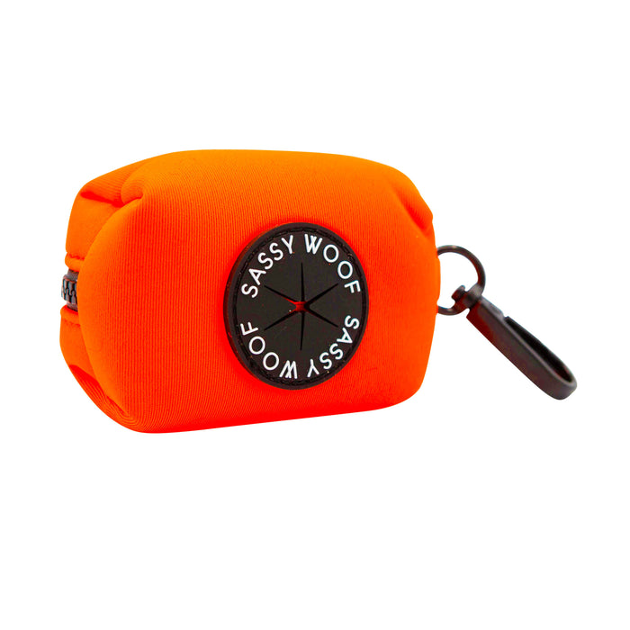 A vibrant neon orange rectangular pouch featuring a black zipper, a circular Sassy Woof logo, and a black clip on one end. This soft pouch is the perfect accessory to pair with the Dog Collar Three Piece Bundle - Neon Orange by Sassy Woof, making it ideal for storing small essentials and complementing any matching dog accessories.