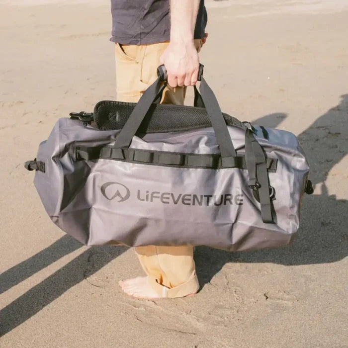 Person holding a Lifeventure Heavy Duty Dry Bag Duffle 60L by the carry handles whilst walking on a beach