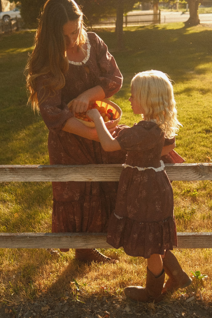 A woman and a young blonde girl stand by a wooden fence in a sunlit grassy field, both wearing matching Ivy City Co Mini Asha Dresses in Warm Cocoa Valencia Toile with charming balloon sleeves, as they share a small basket filled with fresh red and yellow tomatoes.
