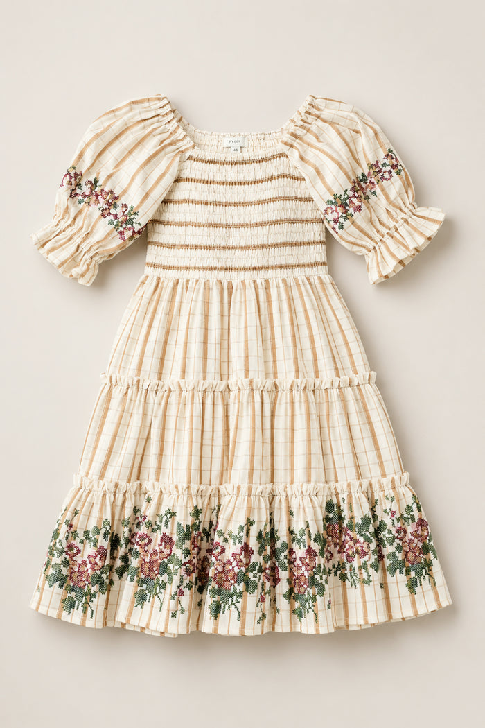 The Mini Madeline Dress in Embroidered Cider Grid by Ivy City Co is a cream-colored, plaid girls’ dress with a smocked bodice, puffed sleeves, and ruffled hem. Charming floral embroidery in green and burgundy decorates the sleeves and skirt.