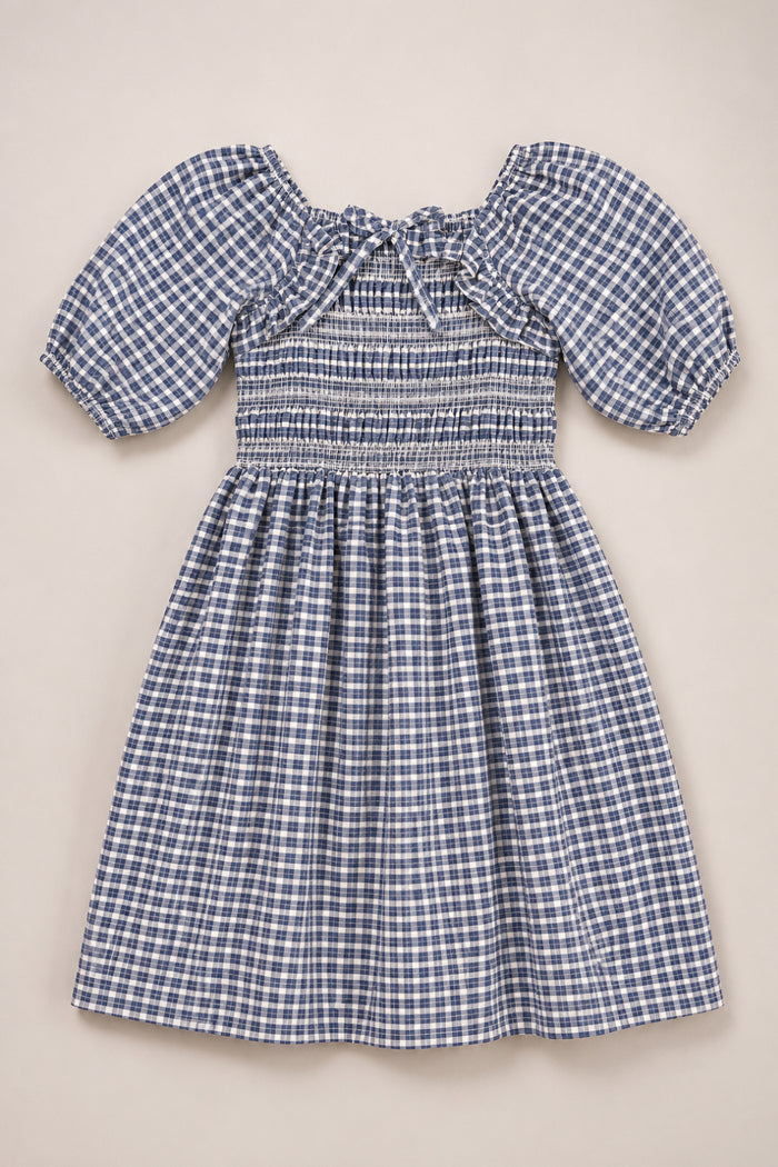 The Ivy City Co Mini Mae 3/4 Sleeve Dress in Prussian Blue Plaid is a girls’ cotton dress featuring blue and white plaid, puffed 3/4 sleeves, a smocked bodice, gathered skirt, and a small bow at the neckline. Shown laid flat on a plain light background.