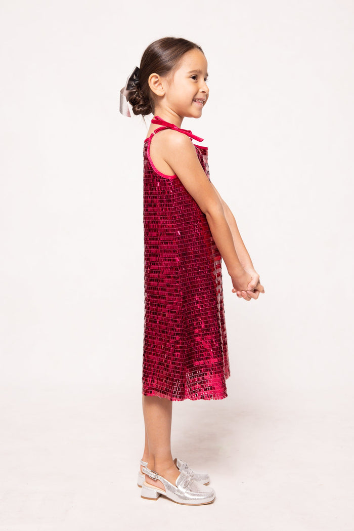 A young girl stands sideways, smiling, dressed in Ivy City Co’s Mini Zelda Dress in Fuchsia Shingle Sequin—a dazzling sleeveless party dress—paired with silver shoes. Her hair is styled in a neat bun and her hands are gracefully clasped in front of her.