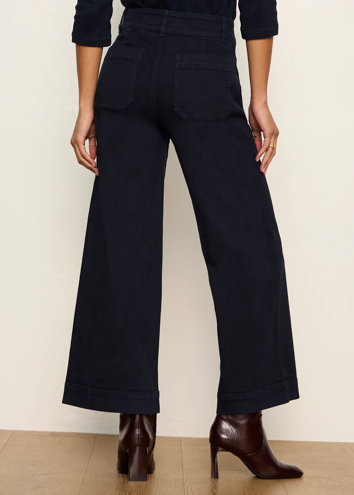 A person shown from the back wearing dark navy wide-leg cropped pants with a cuffed hem, paired with dark brown ankle boots against a plain light beige wall and wood floor.