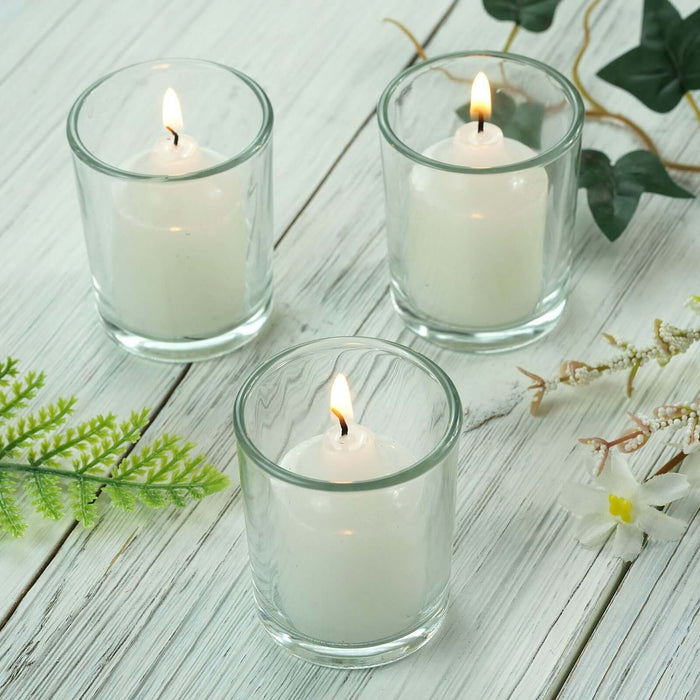 Three white candles in glass holders on a wooden table with greenery accents.