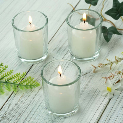 Three white candles in glass holders on a wooden table with greenery accents.
