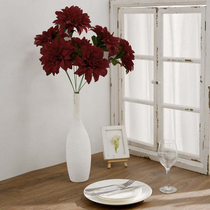 Red artificial flowers in a white vase on a wooden table with glassware.