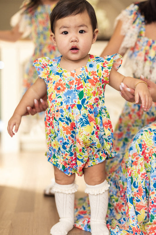 A toddler wears the Ivy City Co Baby Ada Romper in Vibrant Yarrow Print and white knee-high socks, standing indoors supported by an adult in a matching dress. The child looks directly at the camera with a neutral expression.
