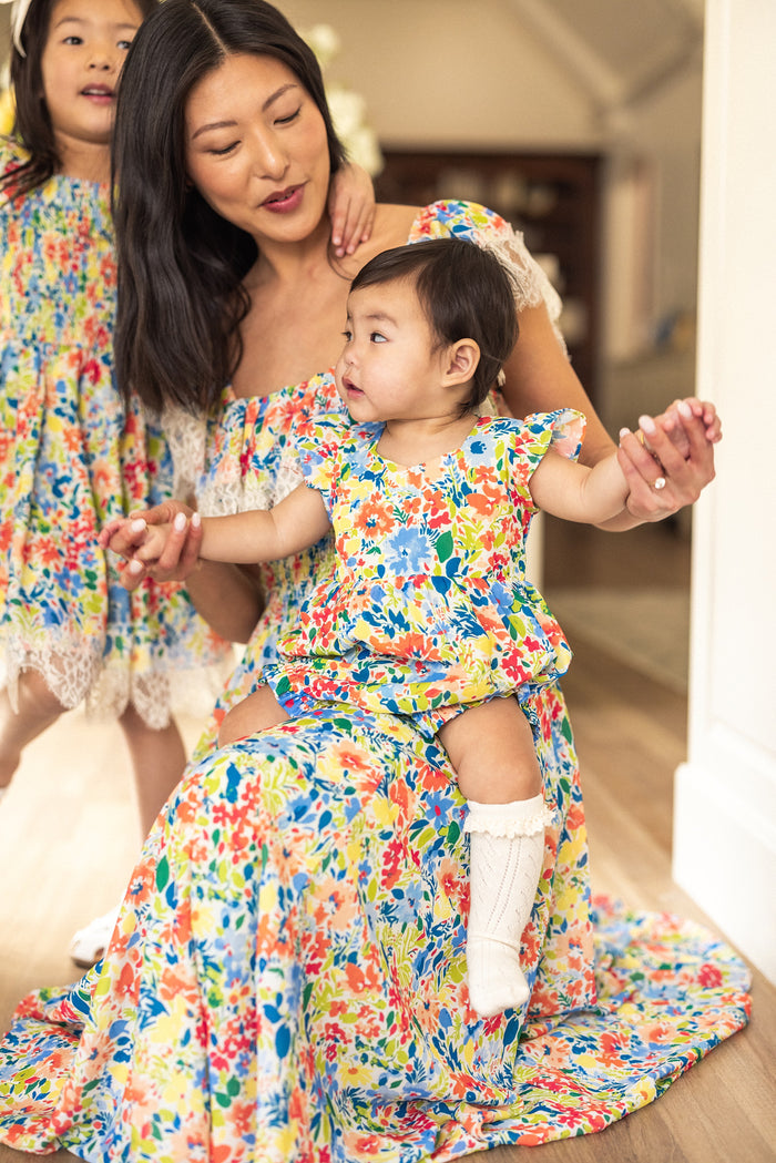 A woman and two children wear matching Ivy City Co outfits in a cheerful indoor scene. The toddler sits on her lap in the Baby Ada Romper in Vibrant Yarrow Print, while another child in a matching dress stands nearby.