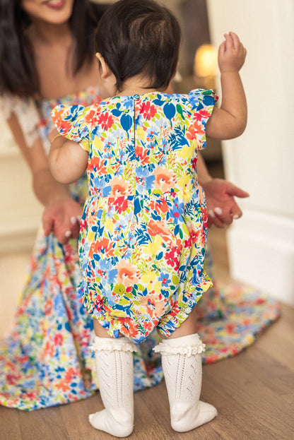 A baby in the Ivy City Co Baby Ada Romper in Vibrant Yarrow Print and white knee-high socks stands facing a smiling woman in a matching Yarrow dress, who holds out her arms to encourage the baby to walk.