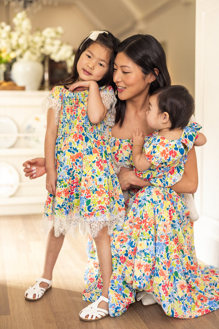 A woman and two young girls wear matching Ivy City Co Mini Ada Dresses in Vibrant Yarrow Print. The older girl stands smiling while the woman kneels, holding the younger girl, all indoors on light wooden floors with a bright background.
