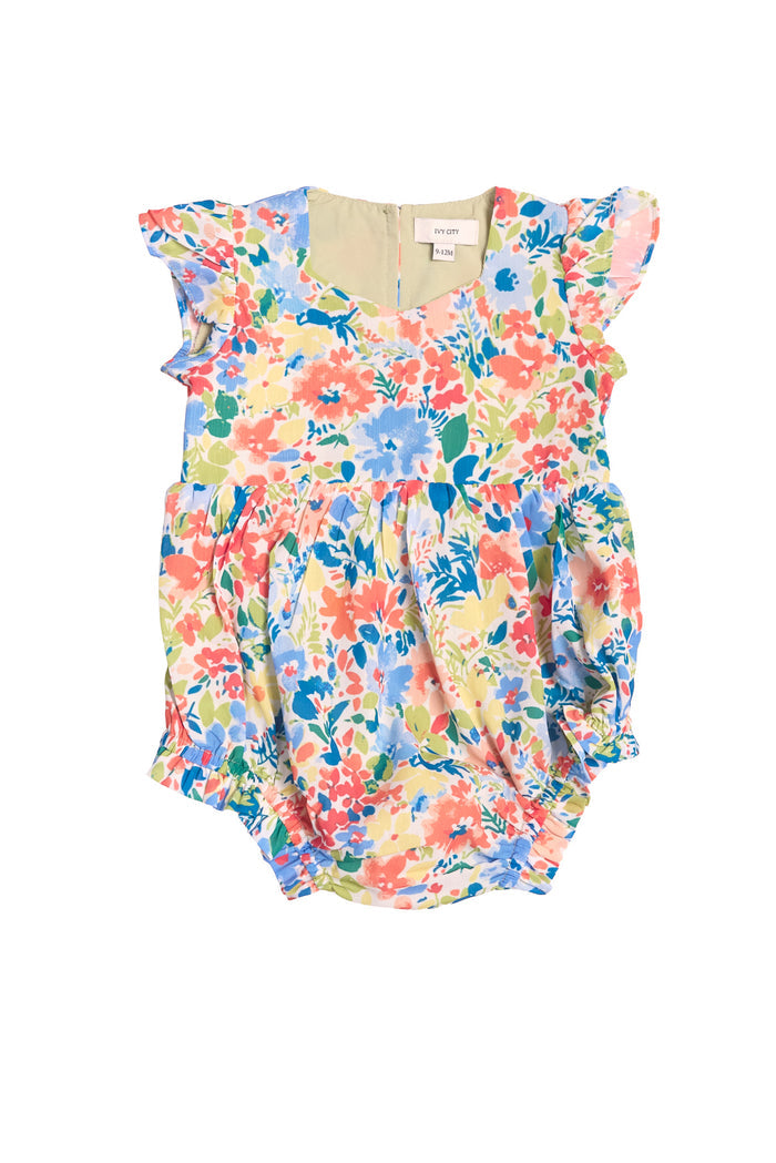 The Ivy City Co Baby Ada Romper in Vibrant Yarrow Print features blue, pink, yellow, and green florals, short ruffled sleeves, gathered leg openings, and soft lightweight fabric.