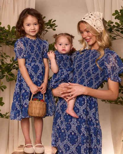 A woman and two girls wear Ivy City Co’s matching blue Trellis Foulard dresses, including the Baby Cordelia Romper. Posing indoors among plants, the older girl holds a small basket for a perfect Easter look.