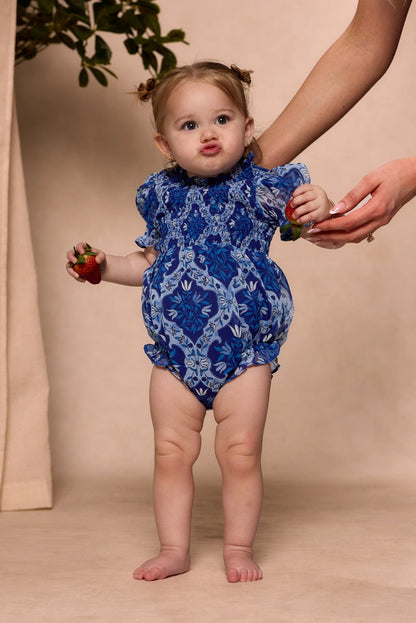 A toddler wearing the Ivy City Co Baby Cordelia Romper in Trellis Foulard, featuring a smocked bodice and blue patterned fabric, stands barefoot holding strawberries in both hands as an adult supports her by the arm. The child makes a playful face with puckered lips.