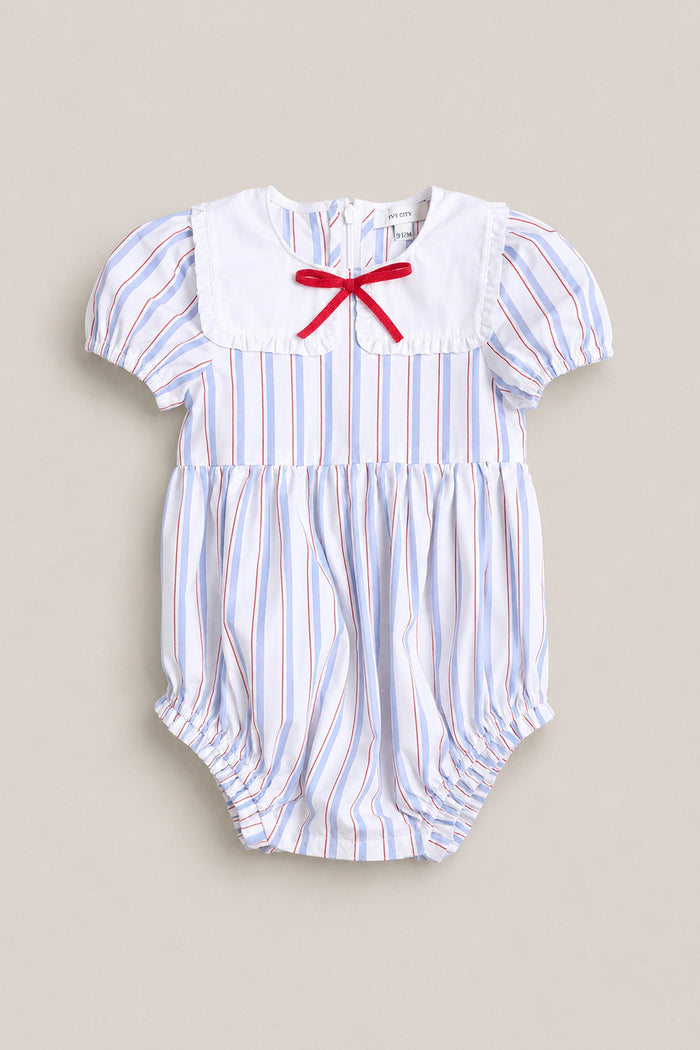 The Ivy City Co Baby Emery Romper in Heritage Stripe features blue and red vertical stripes, a white bib collar with a small red bow, short puff sleeves, and gathered leg openings. Perfect Fourth of July outfit for your little one.