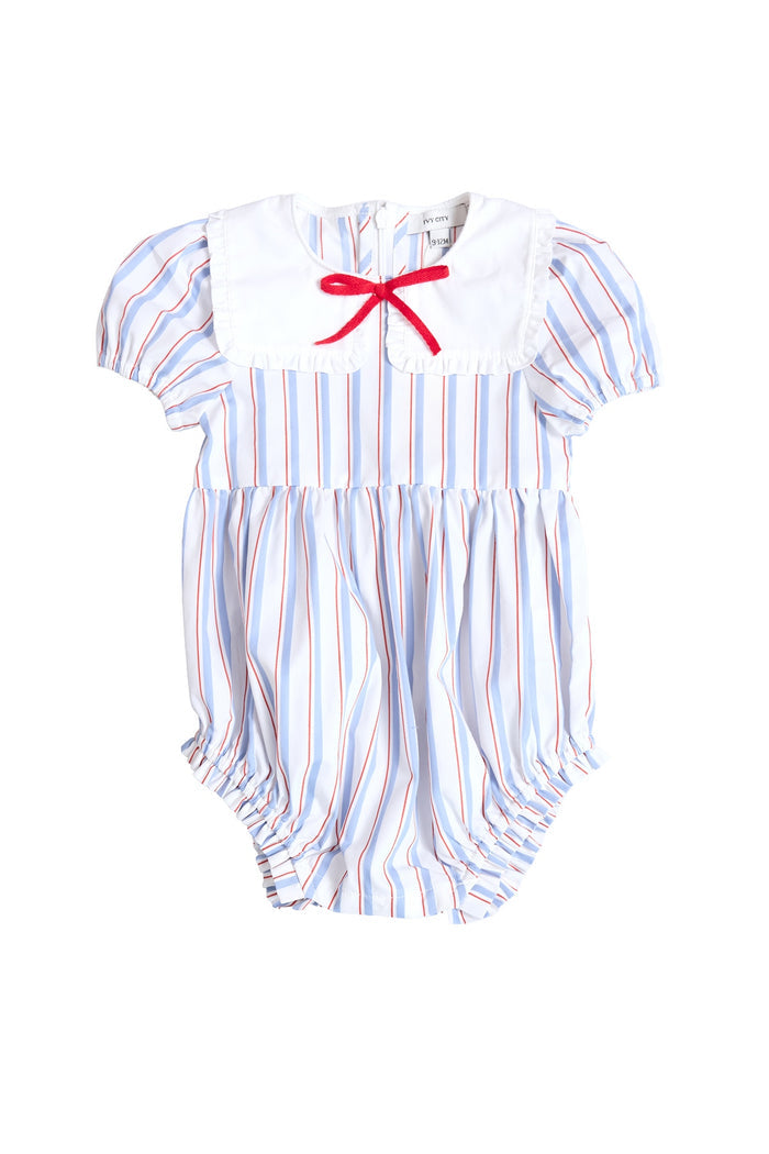 The Ivy City Co Baby Emery Romper in Heritage Stripe features blue, red, and white stripes, puffed short sleeves, and a white bib collar with a red bow—an adorable Fourth of July outfit.