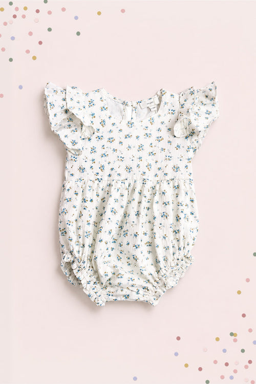 The Ivy City Co Baby Hattie Romper in Madeline Blue features a white cotton fabric with blue floral print, ruffled short sleeves, and elastic leg openings—perfect for special occasions. Shown on a light pink background with colorful confetti dots.