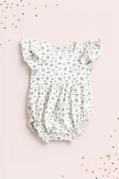 Dress your little one in the Baby Hattie Romper in Madeline Blue by Ivy City Co—crafted from white cotton with a charming blue floral print, ruffled sleeves, and elastic leg openings—ideal for a baby birthday outfit. Photographed against a light pink background adorned with scattered pastel-colored confetti.