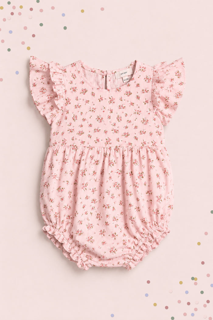 The Ivy City Co Baby Hattie Romper in Madeline Pink features ruffled sleeves and a floral print, displayed on a matching background with pastel confetti—an adorable first birthday outfit.