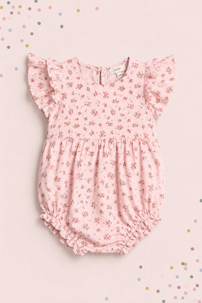 The Ivy City Co Baby Hattie Romper in Madeline Pink features ruffled sleeves and a floral print, displayed on a matching background with pastel confetti—an adorable first birthday outfit.