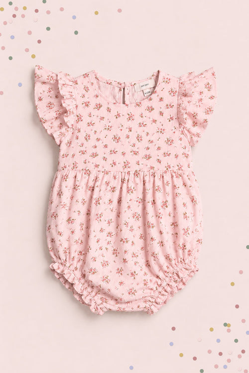 The Ivy City Co Baby Hattie Romper in Madeline Pink features ruffled sleeves and a floral print, displayed on a matching background with pastel confetti—an adorable first birthday outfit.