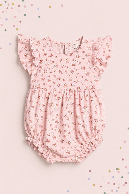 The Ivy City Co Baby Hattie Romper in Madeline Pink features ruffled sleeves and a floral print, displayed on a matching background with pastel confetti—an adorable first birthday outfit.
