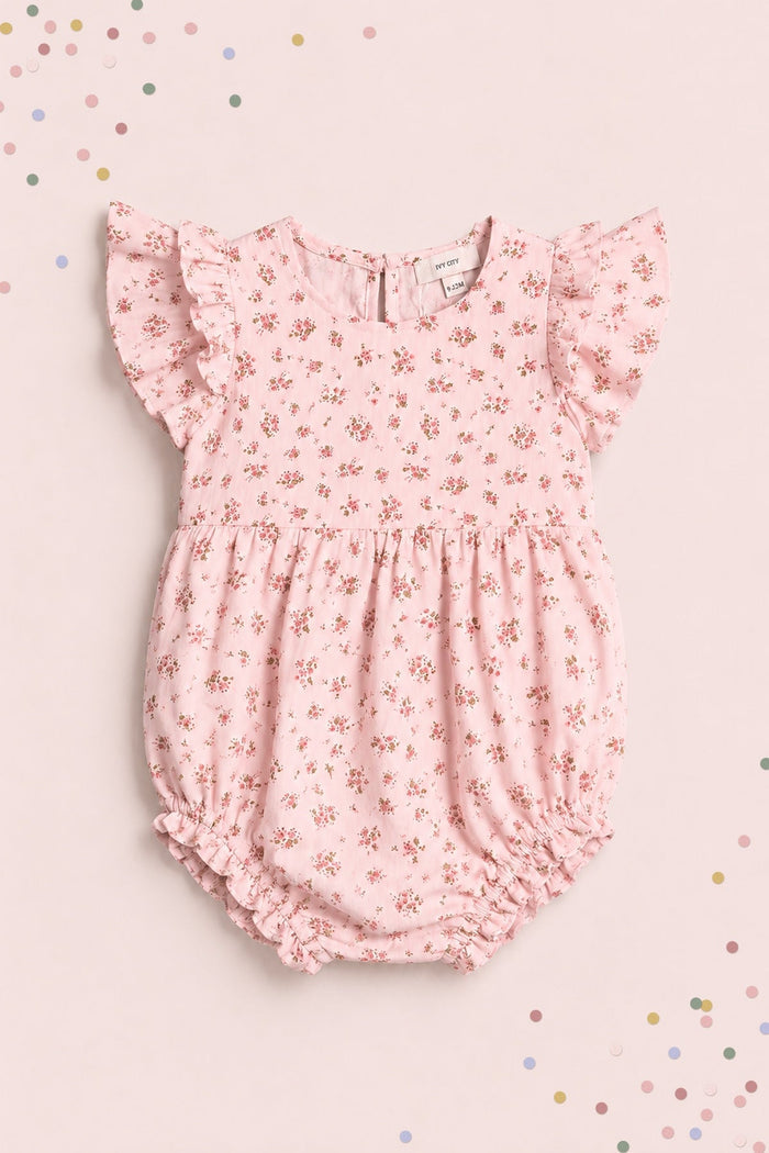 The Ivy City Co Baby Hattie Romper in Madeline Pink is an adorable piece featuring a delicate small floral print, short ruffled sleeves, and elastic leg openings. Shown laid flat on a light pink background with pastel confetti, this charming romper is perfect for your little one.
