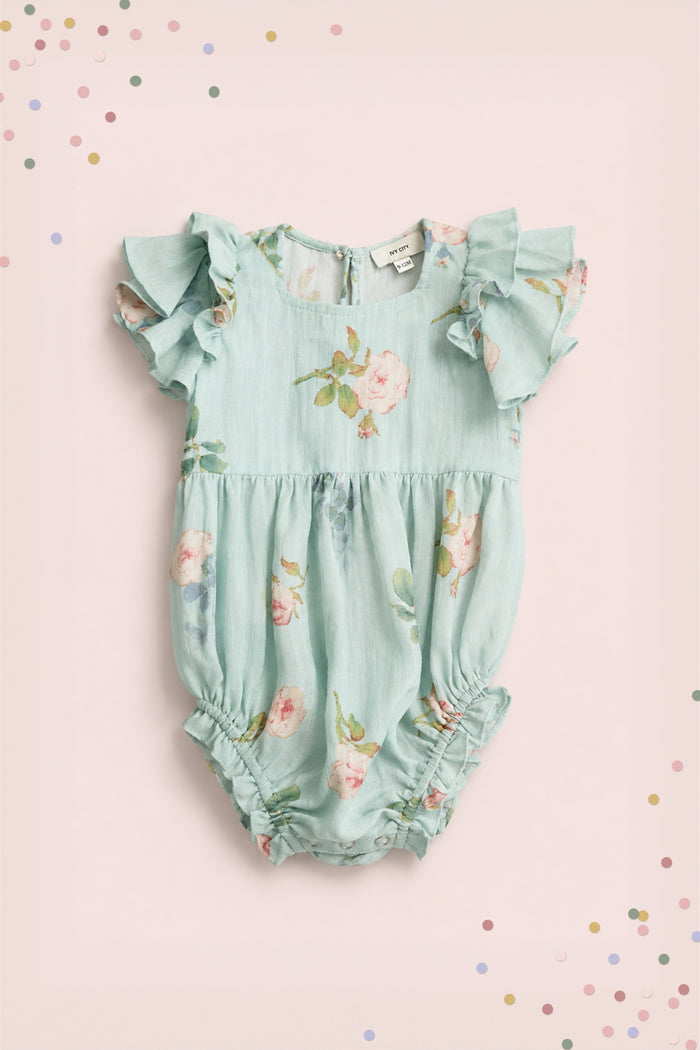 The Ivy City Co Baby Hattie Romper in Roselyn Blue features a light blue cotton-linen fabric with pink floral print, flutter sleeves, and gathered leg openings, pictured on a pale pink background with colorful confetti dots.