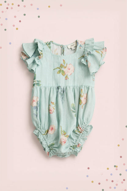 The Ivy City Co Baby Hattie Romper in Roselyn Blue features a light blue cotton-linen fabric with pink floral print, flutter sleeves, and gathered leg openings, pictured on a pale pink background with colorful confetti dots.