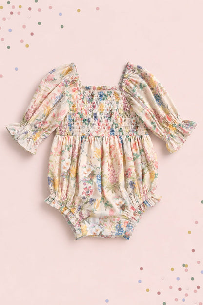 The Baby Madeline Romper in Cordelia Floral by Ivy City Co features a smocked bodice, long puffed sleeves, and charming ruffled cuffs. Shown against a soft pink background with scattered pastel confetti dots, it’s perfect for adorable mommy-and-me moments.