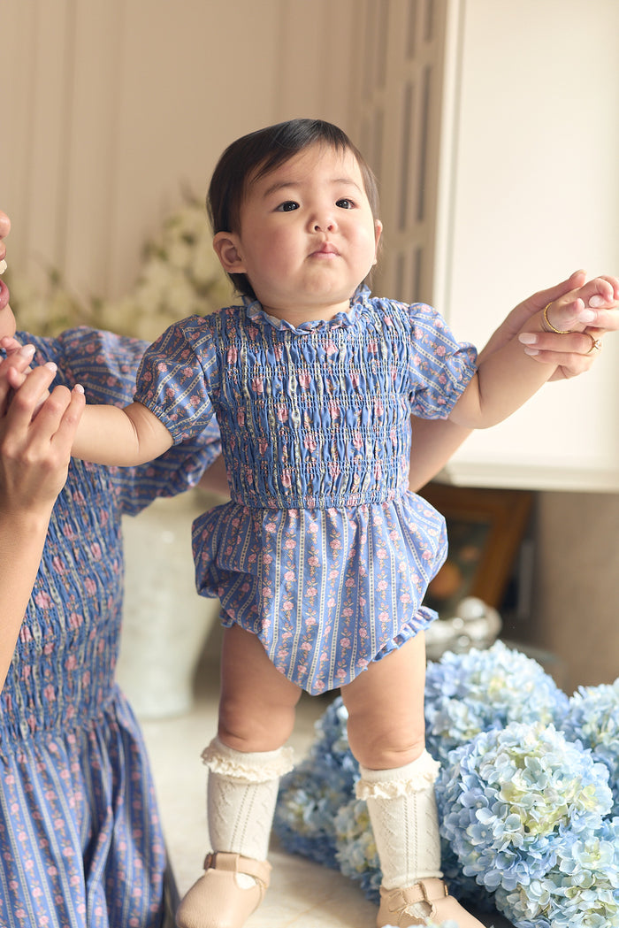 A baby wearing the Ivy City Co Baby Mariah Romper in Linear Print and cream knee-high socks stands, holding an adult's hands. Blue and white hydrangeas bloom in the background as the baby gazes up with curiosity.