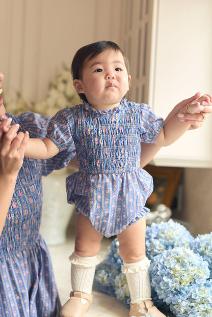 A baby wearing the Ivy City Co Baby Mariah Romper in Linear Print and cream knee-high socks stands, holding an adult's hands. Blue and white hydrangeas bloom in the background as the baby gazes up with curiosity.