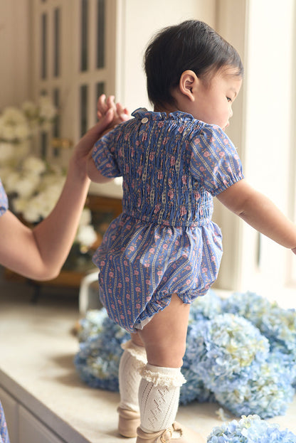 A baby wears the Ivy City Co Baby Mariah Romper in Linear Print with cream knee socks, standing on a windowsill supported by an adult's hand, against a backdrop of blue hydrangeas and white flowers.