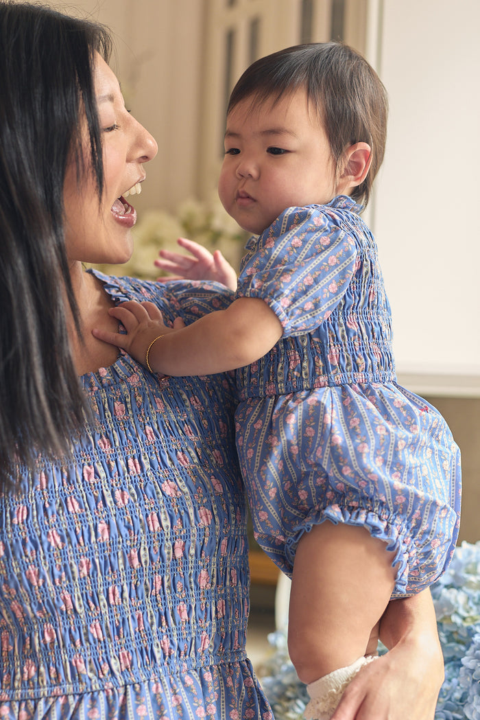 A woman smiles at her baby, both in matching blue outfits with pink patterns. The baby wears the Ivy City Co Baby Mariah Romper in Linear Print and gazes off to the side with a calm expression.