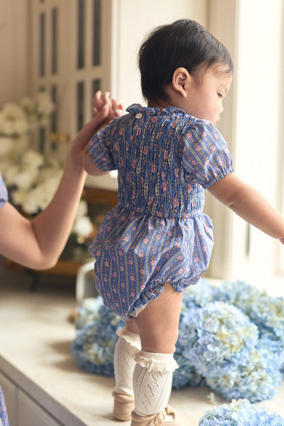 A baby in the Ivy City Co Baby Mariah Romper in Linear Print and cream knee socks stands on a windowsill, holding an adult’s hand, with blue hydrangeas and white flowers in the background.
