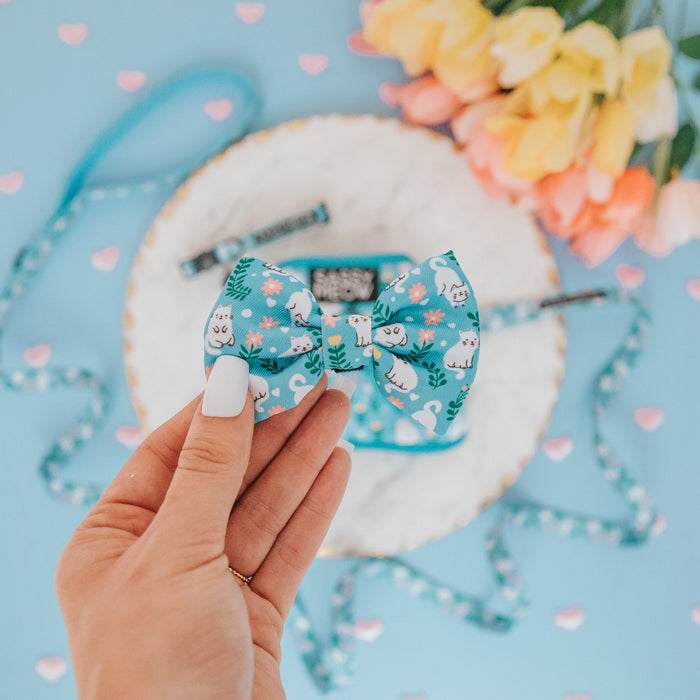 A hand with painted nails holds the Sassy Woof Cat Bowtie - Purrs & Petals, featuring white cat and floral prints. In the background are yellow and pink tulips, a small gift box, decorative ribbons, and heart shapes on a blue surface.