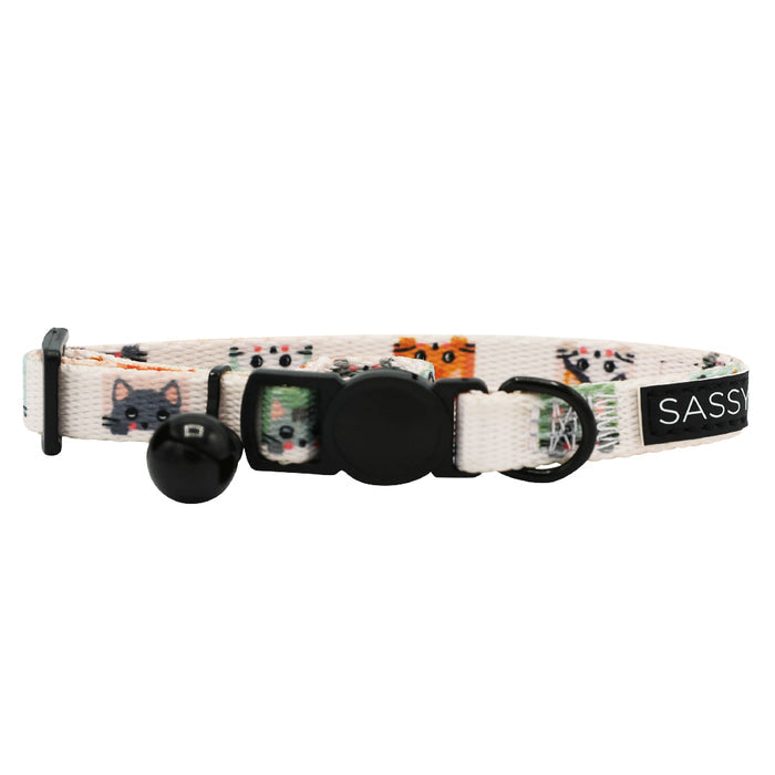 The Cat Collar - Smitten Kittens by Sassy Woof is a white adjustable neoprene collar featuring cartoon cat faces, a black buckle, adjustment slider, metal ring, small bell, and a "SASSY" tag.