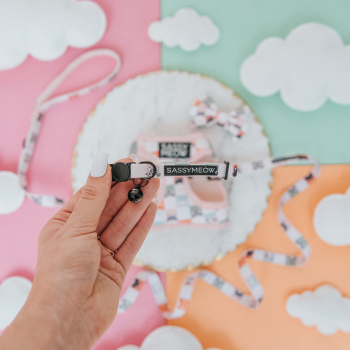 A hand holds a pink Cat Collar - Smitten Kittens by Sassy Woof, featuring a bell and adjustable fit, shown against a pastel cloud background with a coordinating harness and leash set.