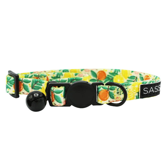 The Sassy Woof Cat Collar - Zest Friends is an adjustable collar with a vibrant tropical fruit pattern, black bell, safety buckle, small black ring, and a stylish SASS label for extra flair.