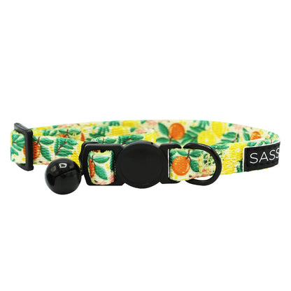 The Sassy Woof Cat Collar - Zest Friends is an adjustable collar with a vibrant tropical fruit pattern, black bell, safety buckle, small black ring, and a stylish SASS label for extra flair.