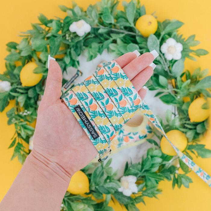 A hand with long, manicured nails holds the Sassy Woof Cat Leash - Zest Friends, featuring a sturdy D-ring and neoprene handle with lemon and leaf designs. A lemon wreath sits behind it on a vibrant yellow surface.