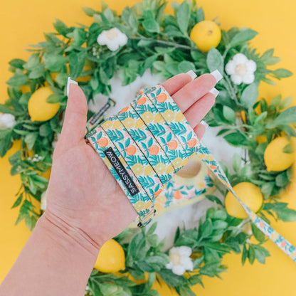 A hand with long, manicured nails holds the Sassy Woof Cat Leash - Zest Friends, featuring a sturdy D-ring and neoprene handle with lemon and leaf designs. A lemon wreath sits behind it on a vibrant yellow surface.