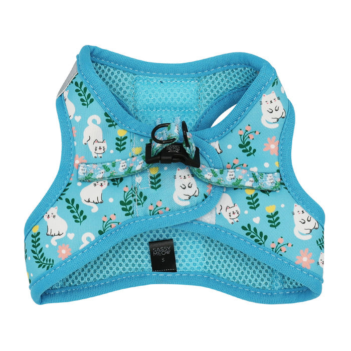 The Sassy Woof Cat Step-In Harness - Purrs & Petals features blue floral fabric with white cats, green leaves, and pink flowers, plus mesh lining, black buckles, an AirTag pocket, and a Sassy Woof S label for secure, stylish comfort.