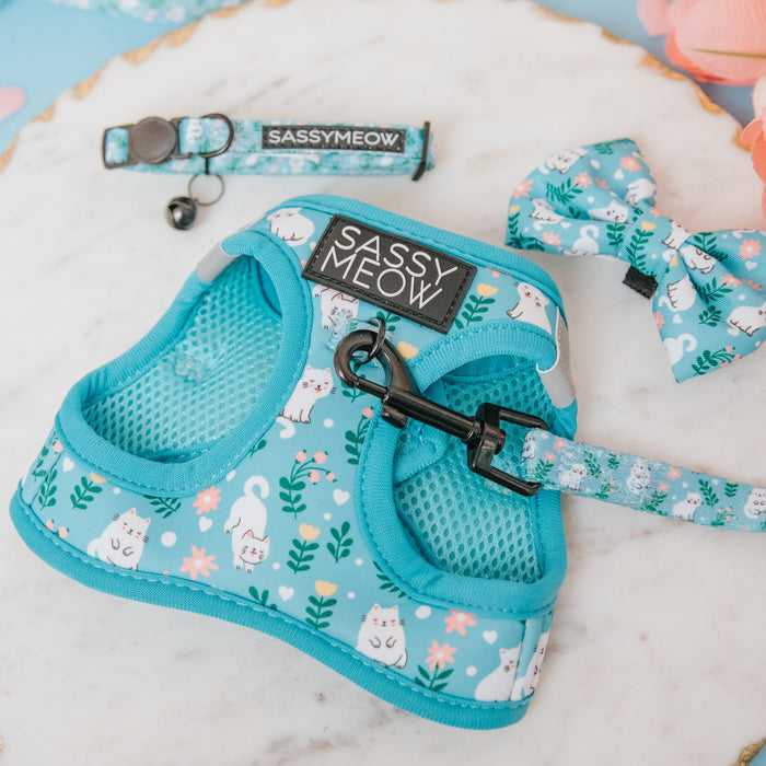 The Sassy Woof Cat Step-In Harness - Purrs & Petals is a blue floral cat harness, leash, collar, and bow tie set with white cat and flower patterns, featuring a custom neoprene harness with an AirTag pocket and SASSYMEOW tag. Displayed on marble.
