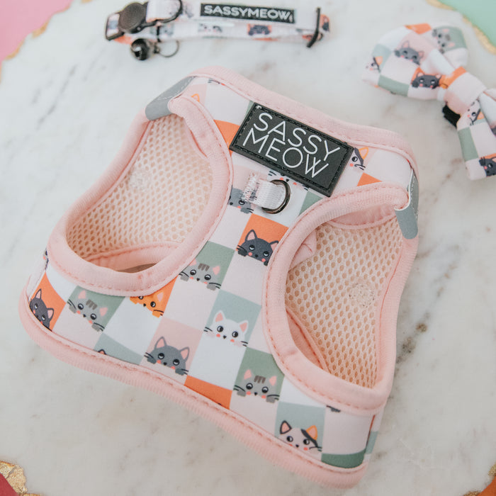 The Sassy Woof Cat Step-In Harness - Smitten Kittens, in pastel pink with a playful pattern and reflective strips, is displayed on marble next to a matching collar and bowtie featuring checkered cartoon cat faces.