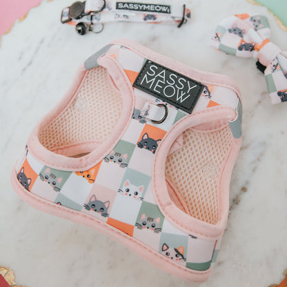 The Sassy Woof Cat Step-In Harness - Smitten Kittens, in pastel pink with a playful pattern and reflective strips, is displayed on marble next to a matching collar and bowtie featuring checkered cartoon cat faces.