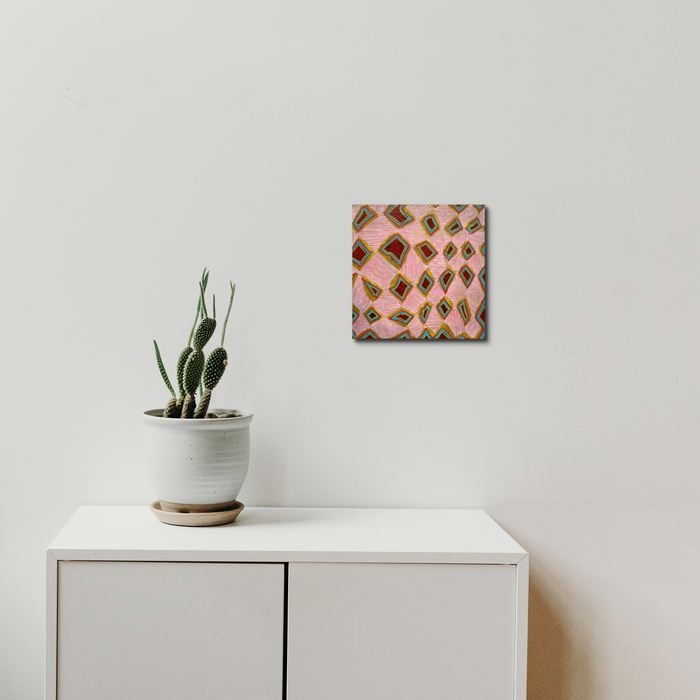 The artwork 'Composition No. 235' by Raul Ortiz features a geometric abstract pattern of repeating diamonds in pink and brown tones on linen canvas, displayed above a white cabinet with a potted cactus. The 12x12 inch acrylic painting creates a rhythmic, modernist design with precise brushwork and balanced composition by Raul Ortiz.