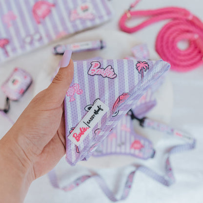 A hand holds the Sassy Woof Dog Bandana - BARBIE™ Closet Goals, a purple and white striped cooling bandana with Barbie-themed designs. Matching Barbie x Sassy Woof pet accessories, like a pink leash, are seen in the background.