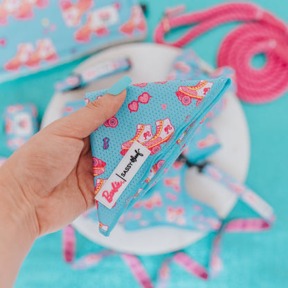 A hand with blue-painted nails holds the Sassy Woof Dog Bandana - BARBIE™ On a Roll, featuring pink roller skates and hearts. The lightweight mesh bandana pops among other matching accessories on a turquoise surface.