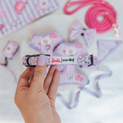 A hand with lavender-painted nails holds the Sassy Woof Dog Collar - BARBIE™ Closet Goals. The “Barbie” and “Sassy Woof” logos are visible, with other matching Sassy Woof pieces and a pink leash blurred in the background.
